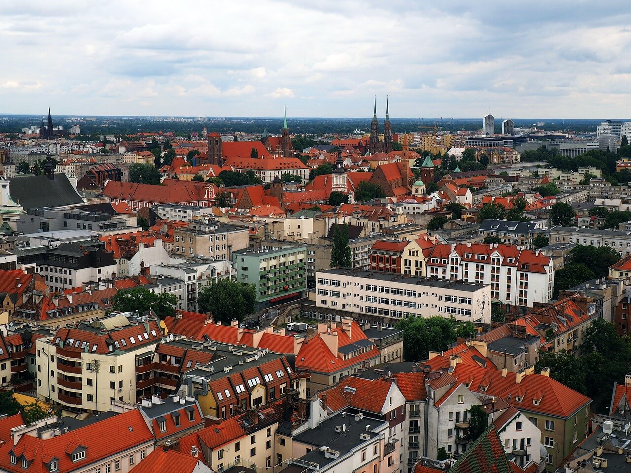 In the picture, we see the panorama of the European city of Wrocław – an aerial view reveals dense buildings with a predominance of red roofs, typical of many historical districts. Among them, the tall towers of churches and historic buildings stand out, giving the place its character.